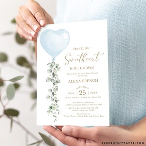 May include: A blue and green baby shower invitation with a heart-shaped balloon and eucalyptus leaves. The text reads "Our Little Sweetheart Is On His Way!" and includes the date, time, and location of the event.  RSVP information and registry details are also included.