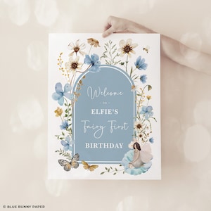 Peut inclure: Une pancarte d'accueil florale bleue et blanche avec le texte "Bienvenue à Elfie's Fairy First Birthday". La pancarte présente une illustration aquarelle de fleurs, de papillons et d'une fée.