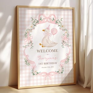 May include: A framed pink and white gingham print with a watercolor illustration of a goose holding a balloon. The text reads "WELCOME to Georgina's 1st Birthday October 25th, 2051". The design includes floral and bow accents.
