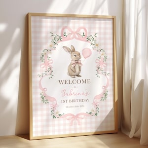 Peut inclure: Une affiche encadr&eacute;e avec un cadre en bois clair. L'impression pr&eacute;sente une illustration &agrave; l'aquarelle d'un lapin tenant un ballon rose, entour&eacute; d'&eacute;l&eacute;ments floraux et de n&oelig;uds. Le texte indique "WELCOME to Sabrina's 1st BIRTHDAY October 25th, 2031."