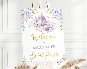 Op de afbeelding: Een welkomstbord voor een bruidsdouche met een paars en gouden bloemenontwerp. Het bord zegt "Welkom bij Savannah's Bridal Shower".
