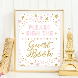 Peut inclure: Une affiche encadrée avec le texte "PLEASE SIGN THE GUEST BOOK" en rose et paillettes dorées. L'affiche est décorée d'étoiles roses et d'accents dorés. Une statue dorée de la Tour Eiffel et une boîte décorative sont à côté de l'affiche.