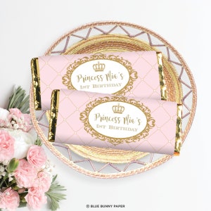 K&ouml;nnte beinhalten: Zwei rosa und goldene Schokoladenriegel-Verpackungen mit einer goldenen Krone und dem Text "Princess Mia's 1st Birthday".
