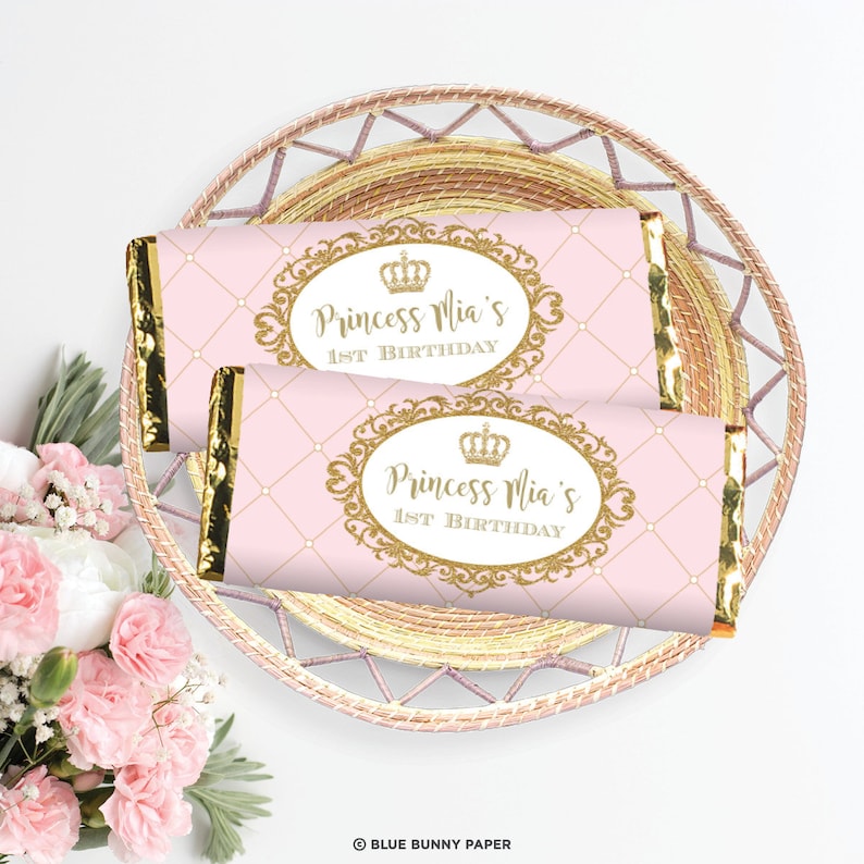 K&ouml;nnte beinhalten: Zwei rosa und goldene Schokoladenriegel-Verpackungen mit einer goldenen Krone und dem Text "Princess Mia's 1st Birthday".