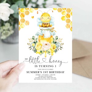 May include: A watercolour invitation card with a bee theme. The card features a cute bee character atop a honey jar, surrounded by flowers and honeycomb patterns. Text reads "Our little honey is turning 1".