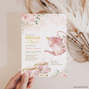 May include: A pink and gold bridal tea party invitation with a teapot pouring tea into two teacups. The invitation features floral designs and gold accents. The text on the invitation reads "Please join us for a Bridal Tea honoring bride-to-be Sarah Cooper Saturday, 18th February 2:00PM - 4:00PM 123 Blush Gold Floral Ave Sydney NSW 2345 RSVP Emily 0412 456 456 by 5th February 20XX Registered at Target, Amazon, and Bed, Bath & Beyond"