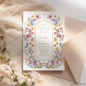 May include: A white floral wedding rehearsal dinner invitation with gold lettering. The invitation features a floral wreath with pink, yellow, and blue flowers. The text reads "Join us for a Rehearsal Dinner honoring Maura and George".