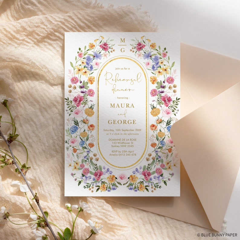 May include: A white floral wedding rehearsal dinner invitation with gold lettering. The invitation features a floral wreath with pink, yellow, and blue flowers. The text reads "Join us for a Rehearsal Dinner honoring Maura and George".