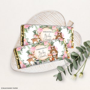 May include: Two white chocolate bar wrappers with a woodland baby shower theme. The wrappers feature a pink floral wreath with a deer, fox, bear, and other woodland creatures. The text on the wrappers reads "Samantha's Baby Shower".