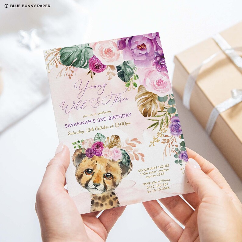 Op de afbeelding: Een roze en paarse bloemen-verjaardagsuitnodiging met een cheeta-welpje dat een bloemenkrans draagt. De uitnodiging zegt "Young Wild & Three" en bevat de tekst "Savannah's 3rd Birthday, Saturday, 12th October at 12:00pm".