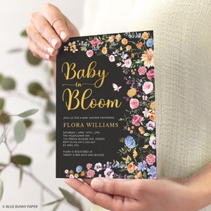 May include: A black baby shower invitation with gold lettering that reads "Baby in Bloom" and floral illustrations. The invitation includes the name Flora Williams, the date and time of the shower, the location, and RSVP information. The text also states that Flora is registered at Target and Bed Bath and Beyond.