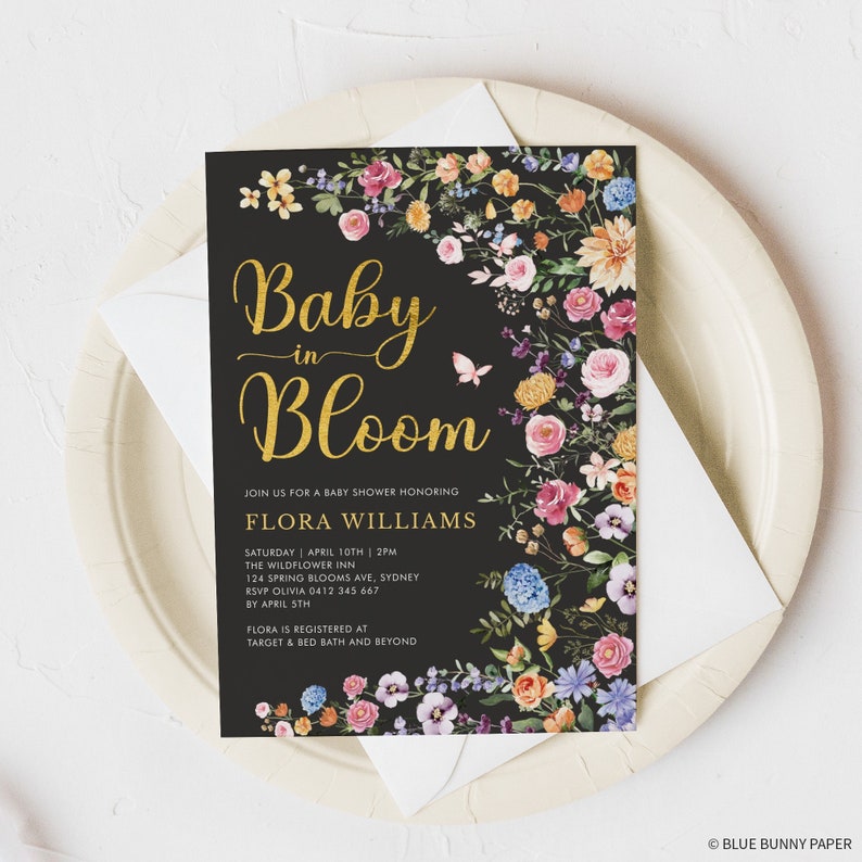 May include: A black baby shower invitation with gold lettering that reads "Baby in Bloom" and floral accents. The invitation includes the name Flora Williams, the date and time of the shower, the location, and RSVP information. It also states that Flora is registered at Target and Bed Bath and Beyond.