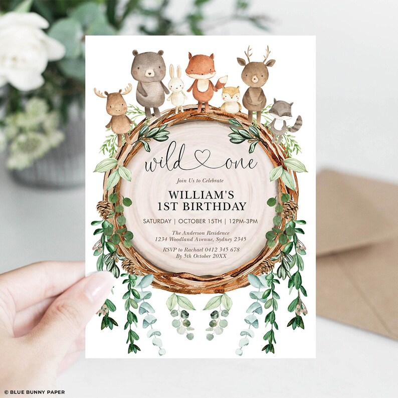 May include: A birthday invitation with a woodland theme. The invitation features watercolor illustrations of forest animals, including a bear, rabbit, fox, deer, and raccoon, surrounding a wooden log. The text reads "wild one" and details for William's 1st birthday.