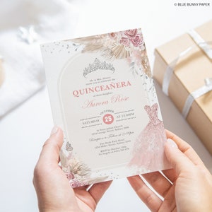 May include: A pink and white Quincea&ntilde;era invitation with a floral design and a crown. The invitation includes the text "Quincea&ntilde;era" and "Aurora Rose" in a pink script font. The invitation also includes the date and time of the event, as well as the location of the church and reception.