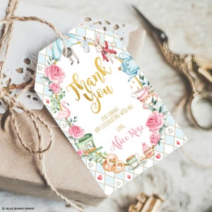 May include: A white thank you tag with a gold border and text. The tag features a watercolor illustration of Alice in Wonderland characters and objects, including a flamingo, a Mad Hatter, a teapot, and a cupcake. The text reads "Thank You" in gold lettering and "For coming and celebrating with me! Love, Alice Rose".