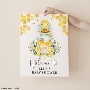 Peut inclure: Une pancarte de bienvenue pour une fête prénatale jaune et blanche avec une abeille de dessin animé assise sur un pot de miel. La pancarte indique "Bienvenue à la fête prénatale d'Ella".