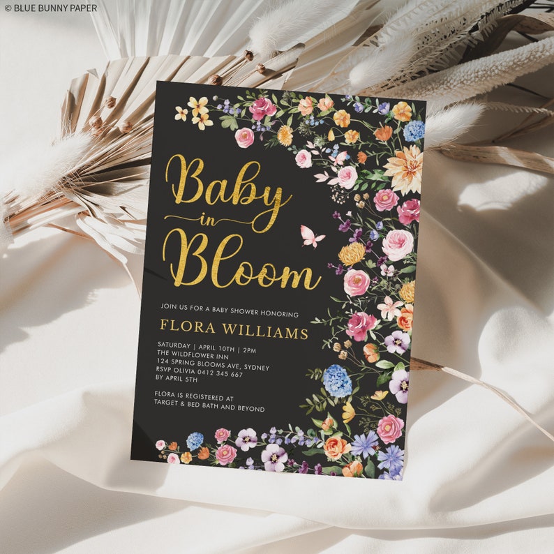 May include: A black baby shower invitation with gold lettering that reads "Baby in Bloom". The invitation features a floral border with pink, yellow, blue, and purple flowers on a black background. The text includes the name Flora Williams, the date and time of the shower, the location, and RSVP information.