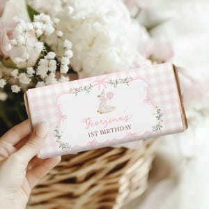 Könnte beinhalten: Schokoladenriegelverpackung mit rosa-weißem Karomuster und dekorativem Etikett. Das Etikett zeigt eine Cartoon-Gans mit einem Ballon, mit dem Text "Georgina's 1st Birthday" in Schreibschrift. Die Verpackung wird vor einem Blumenarrangement gehalten.