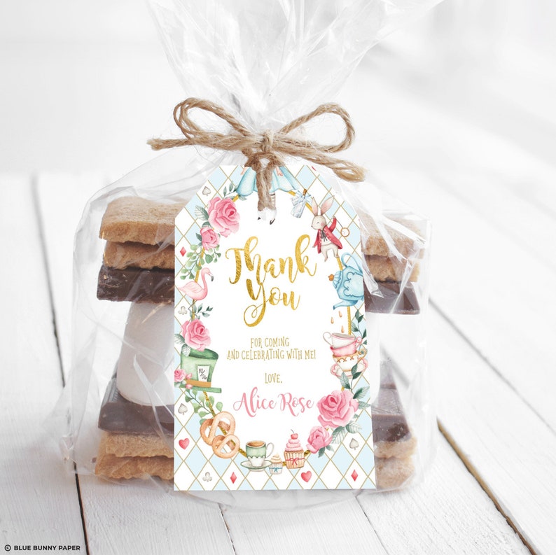 May include: A white thank you tag with a blue and gold Alice in Wonderland theme. The tag features a pink floral wreath with a flamingo, a teapot, a teacup, a Mad Hatter, a White Rabbit, and a playing card. The text on the tag reads "Thank You" in gold lettering and "For coming and celebrating with me! Love, Alice Rose".