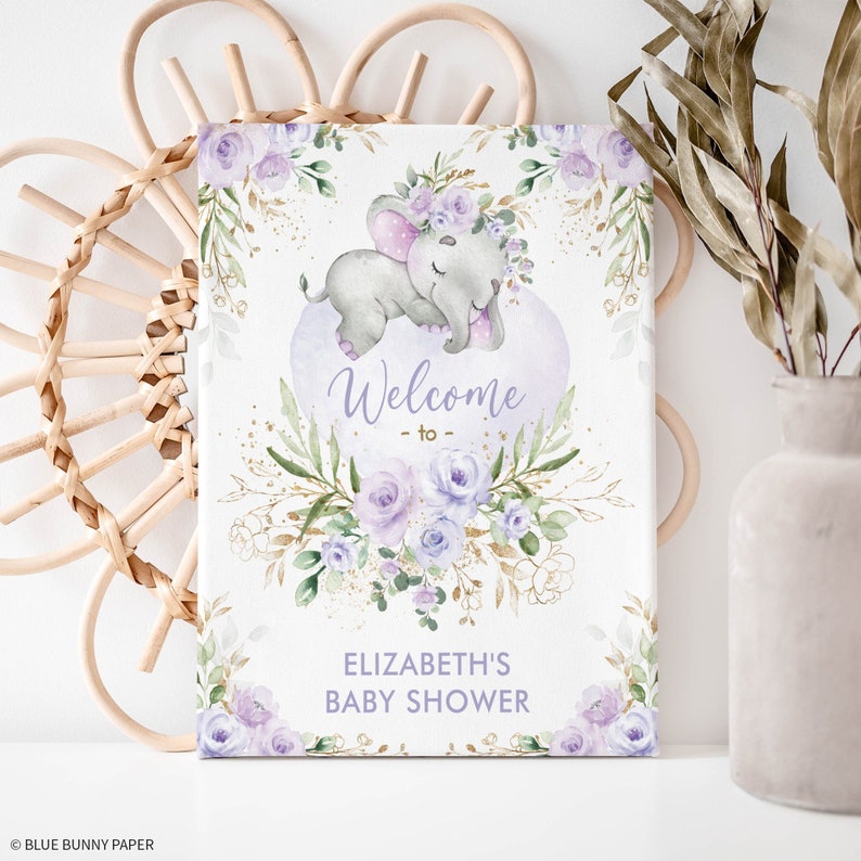 Puede incluir: Un cartel de bienvenida para una baby shower con flores moradas y doradas y una ilustraci&oacute;n de acuarela de un elefante durmiendo con una corona de flores.
