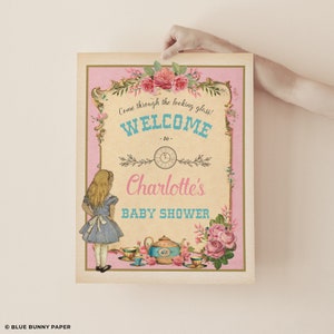 Może przedstawiać: Winietkowy szyld w stylu vintage na baby shower z r&oacute;żowym tłem i złotymi akcentami. Na szyldzie znajduje się kresk&oacute;wkowa postać Alicji z Krainy Czar&oacute;w, czajnik i filiżanki do herbaty. Tekst na szyldzie brzmi "Przejdź przez lustro! Witamy na baby shower Charlotte".