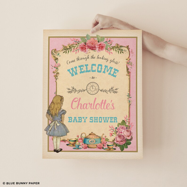 Może przedstawiać: Winietkowy szyld w stylu vintage na baby shower z r&oacute;żowym tłem i złotymi akcentami. Na szyldzie znajduje się kresk&oacute;wkowa postać Alicji z Krainy Czar&oacute;w, czajnik i filiżanki do herbaty. Tekst na szyldzie brzmi "Przejdź przez lustro! Witamy na baby shower Charlotte".
