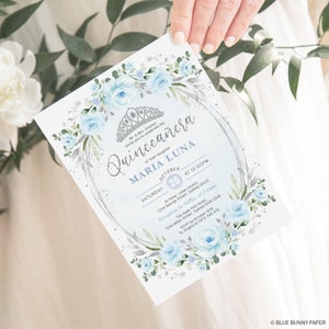 May include: A blue and silver Quincea&ntilde;era invitation with a floral wreath and a crown. The text reads "Mr. & Mrs. Alvarez invite you to celebrate the Quincea&ntilde;era of their daughter Maria Luna, Saturday, October 25th at 12:00pm, St. Peter Julian Church, 1234 George Street, Sydney 2000. Reception to follow at 2:00pm, The Hyde Park Hotel, 1234 Miller Street, Sydney NSW 2345. RSVP by October 10th to Angela at 0412 345 678. Blue Bunny Paper."