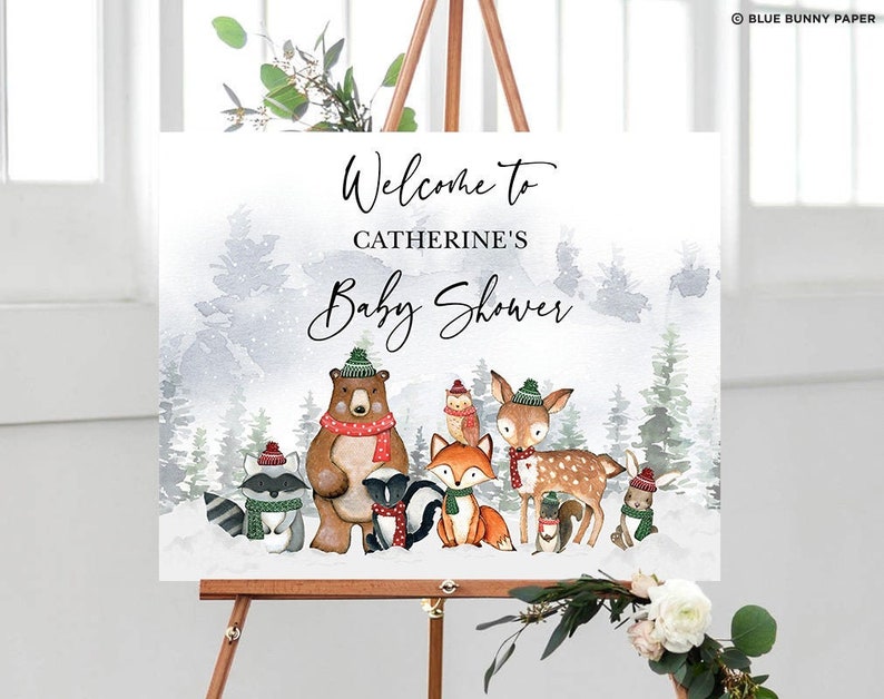 May include: Welcome to Catherine's Baby Shower sign with a winter woodland theme. The sign features a watercolor background with a brown bear, a deer, a fox, a raccoon, a skunk, and an owl. The animals are wearing winter hats and scarves.