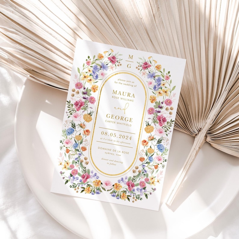 Wildflower wedding invitation with watercolor floral border in pink yellow purple, gold oval frame, on white plate with dried palm leaves.