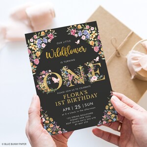 May include: A black and gold floral first birthday invitation for a girl named Flora. The invitation features a gold 'ONE' with floral accents and a butterfly. The text reads 'Our little Wildflower is turning ONE. Please join us to celebrate Flora's 1st Birthday. APR 25 | SUN at 2 o'clock in the afternoon. 123 Spring Boulevard Sydney NSW 2034. RSVP Olivia 0412 345 678 by May 5th.'