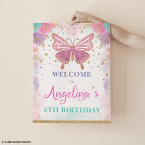 May include: A pink and gold welcome sign with a butterfly design. The sign says "Welcome to Angelina's 5th Birthday".
