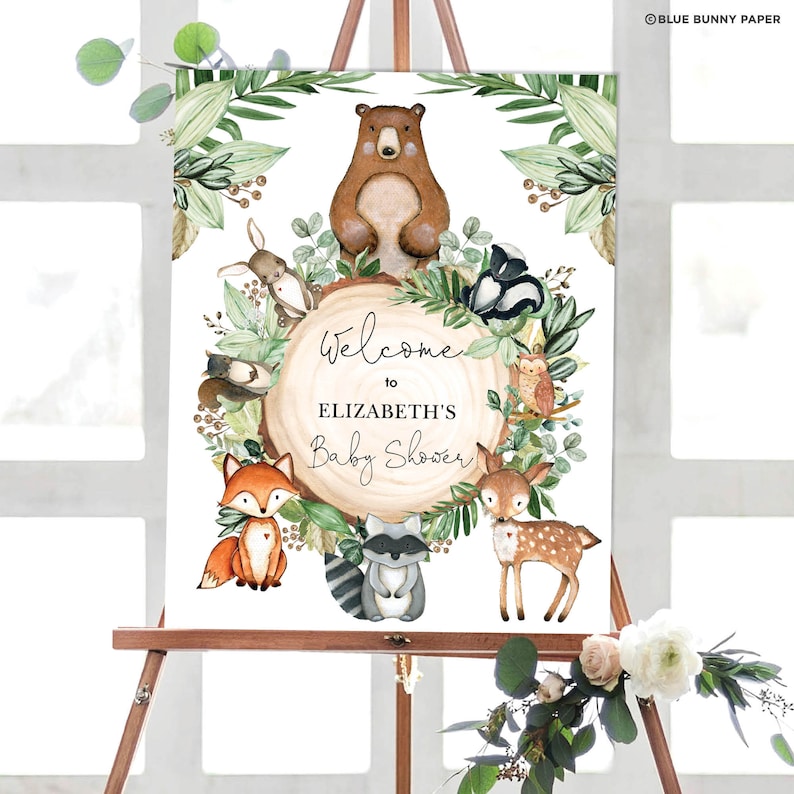 May include: Welcome to Elizabeth's Baby Shower sign with a woodland theme. The sign features a brown bear, a fox, a deer, a raccoon, a rabbit, a skunk, and an owl surrounded by green leaves and a wooden circle with the text "Welcome to Elizabeth's Baby Shower".