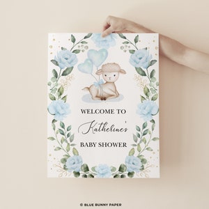 May include: A welcome sign for a baby shower with a cute watercolor illustration of a lamb holding blue balloons. The sign has a floral wreath with blue flowers and gold accents. The text on the sign reads "Welcome to Katherine's Baby Shower".