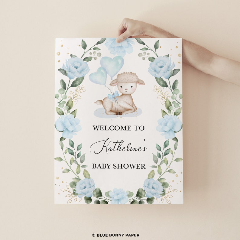 May include: A welcome sign for a baby shower with a cute watercolor illustration of a lamb holding blue balloons. The sign has a floral wreath with blue flowers and gold accents. The text on the sign reads "Welcome to Katherine's Baby Shower".
