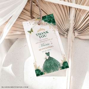 May include: A white tag with a gold-trimmed arch, butterfly, and floral accents. The tag reads "Thank You" and "Maria's Quinceanera 15.05.2031" with a green dress illustration. A brown string is attached to the tag.