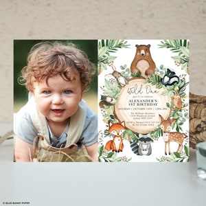 May include: A photo of a smiling baby next to a "Wild One" birthday invitation. The invitation features woodland animals like a bear, fox, and deer, with text that reads "Alexander's 1st Birthday" and event details. The invitation is surrounded by green foliage.