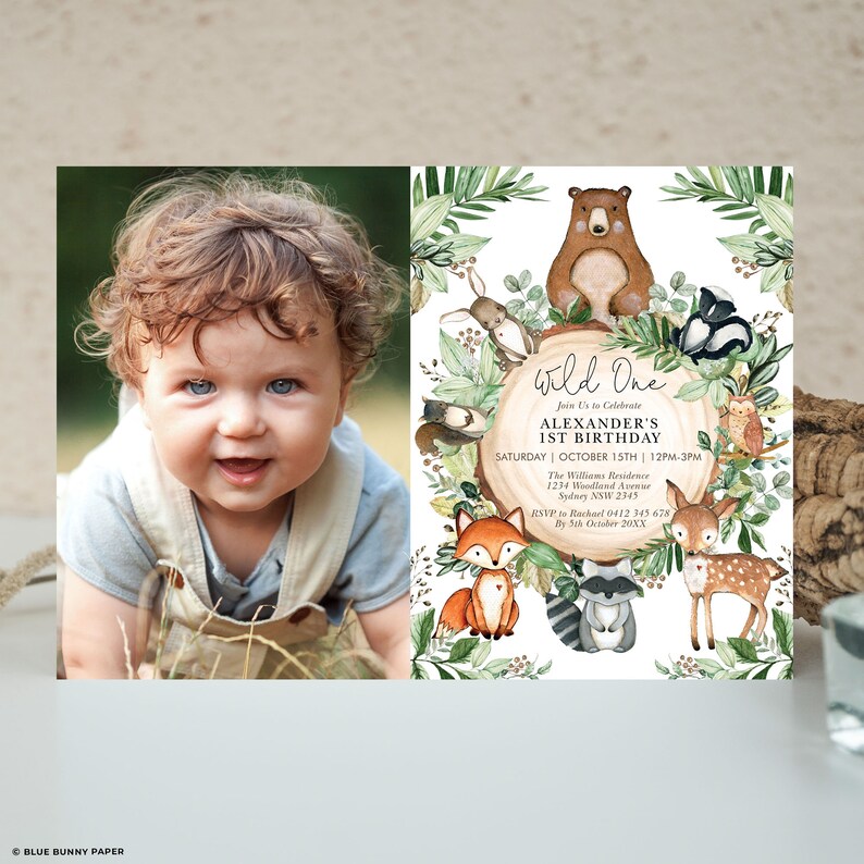 May include: A photo of a smiling baby next to a "Wild One" birthday invitation. The invitation features woodland animals like a bear, fox, and deer, with text that reads "Alexander's 1st Birthday" and event details. The invitation is surrounded by green foliage.