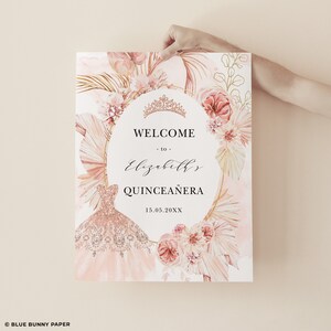 May include: A pink and gold welcome sign for a Quincea&ntilde;era. The sign features a watercolor illustration of a pink dress, flowers, and palm leaves. The text reads "Welcome to Elizabeth's Quincea&ntilde;era 15.05.20XX".