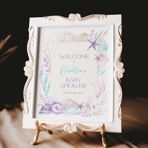 May include: A framed baby shower sign with a white ornate frame and a gold stand. The sign reads "Welcome to Cordella's Baby Shower" with watercolor illustrations of seashells, starfish, and coral in pastel colours. The date is 25th October 2031.