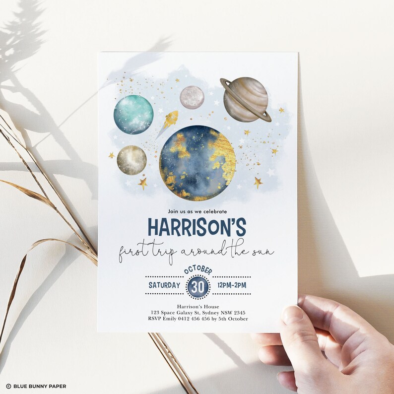 May include: A blue and gold space-themed invitation for a first birthday party. The invitation features a watercolor illustration of planets and stars. The text reads "Join us as we celebrate HARRISON'S first trip around the sun. October 30, Saturday, 12PM-2PM. Harrison's House, 123 Space Galaxy St, Sydney NSW 2345. RSVP Emily 0412 456 456 by 5th October."