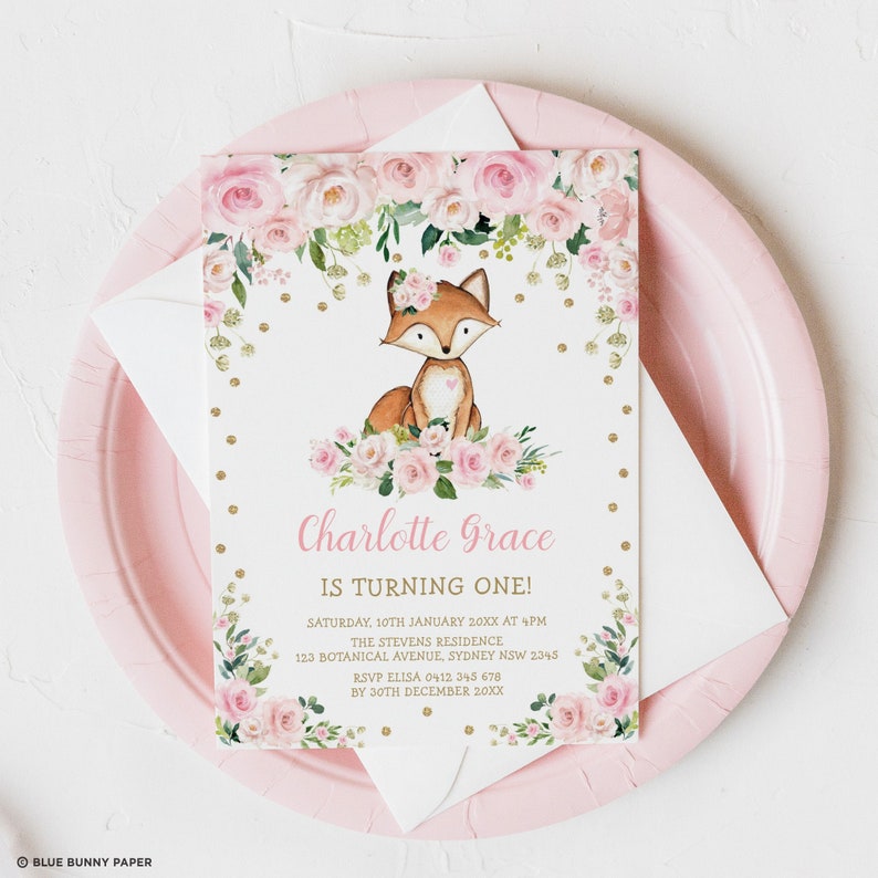 May include: A pink paper plate holds a white envelope and a party invitation. The invitation features a watercolor illustration of a fox with a floral crown and a heart. Text reads "Charlotte Grace is turning one!" with event details.