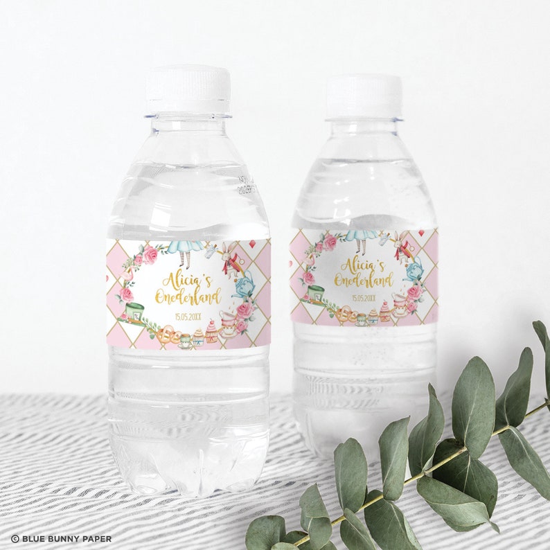 K&ouml;nnte beinhalten: Zwei durchsichtige Plastikwasserflaschen mit rosa und goldenen Etiketten. Die Etiketten zeigen ein fantasievolles Alice-im-Wunderland-Motiv mit Illustrationen von Alice, dem Hutmacher und anderen Figuren. Der Text auf den Etiketten lautet "Alicia's Wonderland" und "15.05.2021".