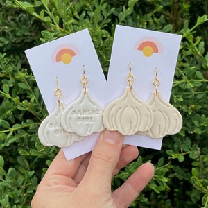 May include: Off-white garlic-shaped earrings with gold-colored hooks. The earrings are displayed on white cards with a rainbow design. One earring has the text "GARLIC GIRL". The background is green foliage.