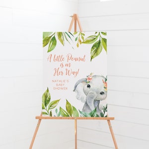May include: A baby shower sign on a wooden easel. The sign features an illustration of a gray elephant with a floral crown and the text "A Little Peanut is on Her Way!" surrounded by green leaves.