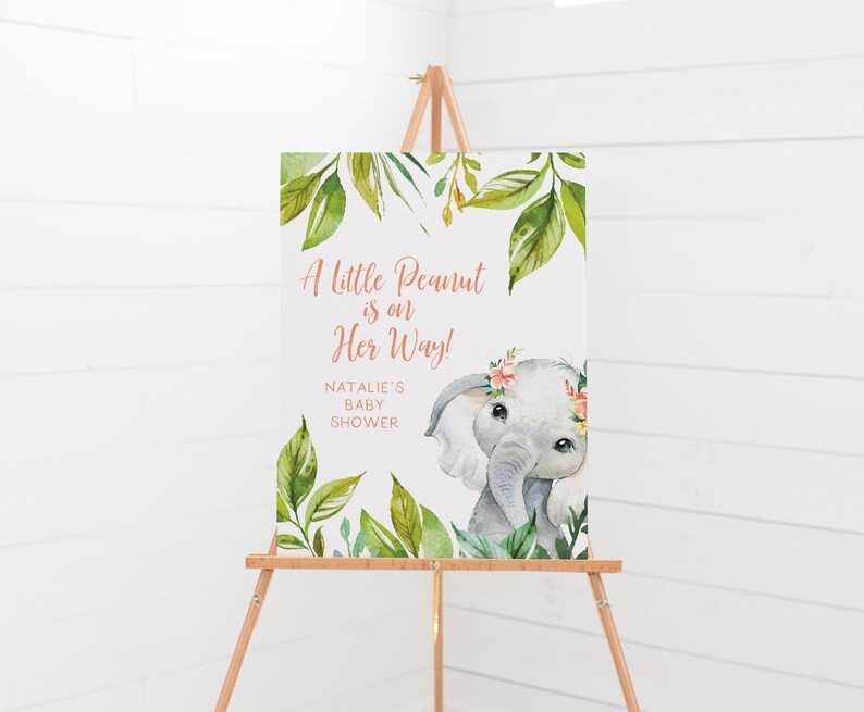 May include: A baby shower sign on a wooden easel. The sign features an illustration of a gray elephant with a floral crown and the text "A Little Peanut is on Her Way!" surrounded by green leaves.