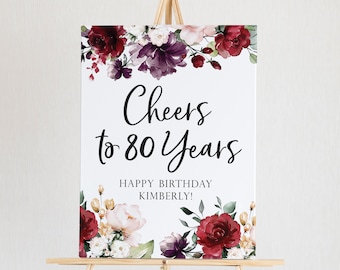 May include: A white sign on a wooden easel with the text "Cheers to 80 Years" and "Happy Birthday Kimberly!". The sign is decorated with burgundy, purple, and cream-colored floral arrangements.