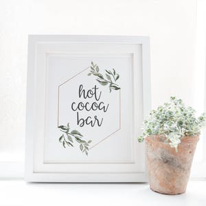 May include: White framed sign with a gold geometric shape and green foliage. The sign reads "hot cocoa bar" in black cursive lettering.