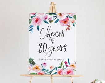 May include: A white sign with a floral border and the text "Cheers to 80 years" in black script. The sign also says "Happy Birthday Wendy!" in black text.