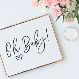 May include: A white framed sign with gold trim, featuring the text "Oh Baby!" in black cursive script with a small heart below the text.