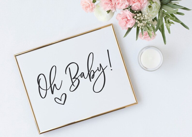 May include: A white framed sign with gold trim, featuring the text "Oh Baby!" in black cursive script with a small heart below the text.